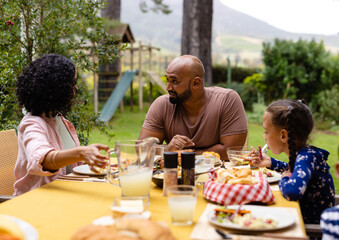 Happy biracial parents, son and daughter sitting at table having meal and talking in garden