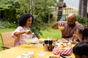 Happy biracial parents, son and daughter sitting at table eating meal in garden, copy space