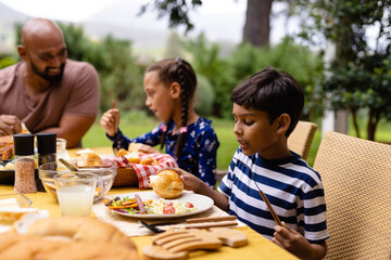 Happy biracial father, son and daughter sitting at table having meal and talking in garden