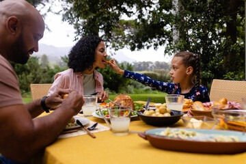 Happy biracial parents and daughter at table having meal and tasting each other's food in garden