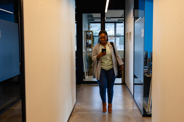 Fototapeta na wymiar Happy plus size african american casual businesswoman using smartphone in office corridor