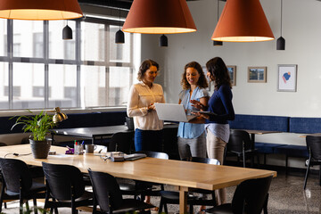 Happy diverse casual businesswomen discussing using laptop and tablet in office