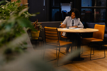 Biracial casual businesswoman taking notes in notebook at table in office lounge
