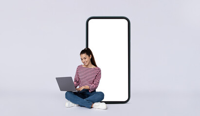 Positive young woman independent contractor sitting by phone using laptop