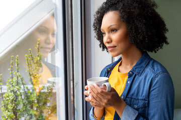 Happy biracial woman holding cup of coffee and looking out window at home