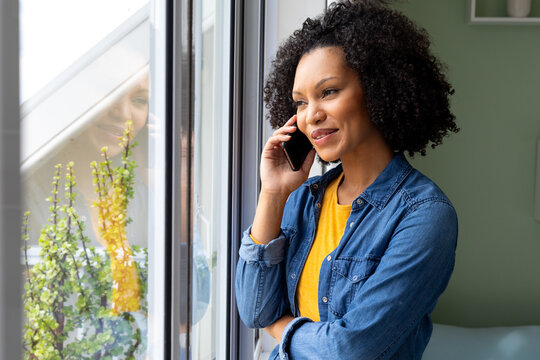 Happy biracial woman talking on smartphone and looking out window at home - Powered by Adobe