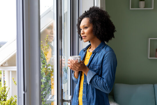 Biracial woman holding cup of coffee and looking out window at home