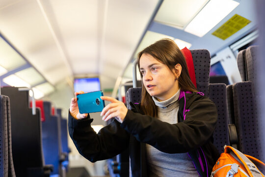 Interested female traveler sitting near window on comfortable seat in express train, filming picturesque mountain landscapes of Switzerland behind glass