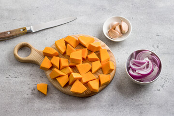 Ingredients for a vegetable dish: chopped pumpkin on a wooden board, chopped red onion, garlic in husk on gray textured background
