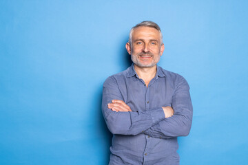 Handsome middle aged grey haired man standing with arms folded isolated on blue background.