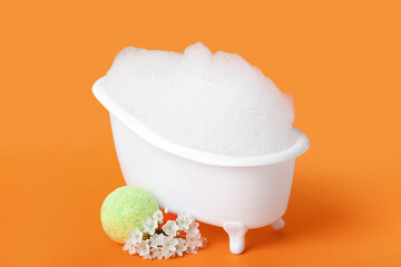 Small bathtub with foam, flowers and bath bomb on color background