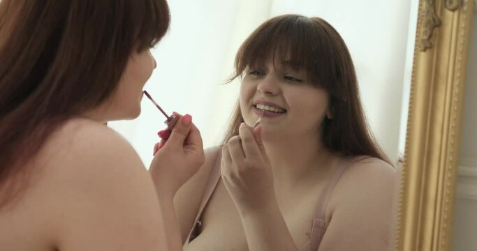 A plus-size Caucasian woman takes center stage near the mirror, delicately painting her lips with vibrant lipstick. With each stroke, she embraces her individuality, celebrating her unique features.