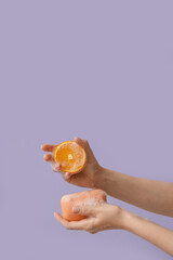 Hands with soap and orange on lilac background