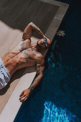Chico joven tatuado y musculoso posando con bañador urbano en piscina de hotel un día soleado