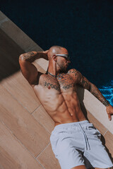 Chico joven tatuado y musculoso posando con bañador urbano en piscina de hotel un día soleado