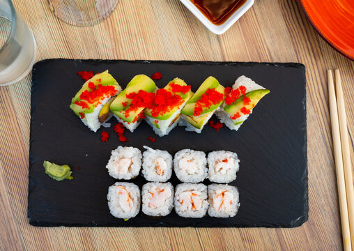 Delicious sushi rolls uramaki filled with salmon, mango and cucumber topped with avocado and tobiko roe and tiger prawn norimaki served on slate board with wasabi and soy sauce. Japanese snack