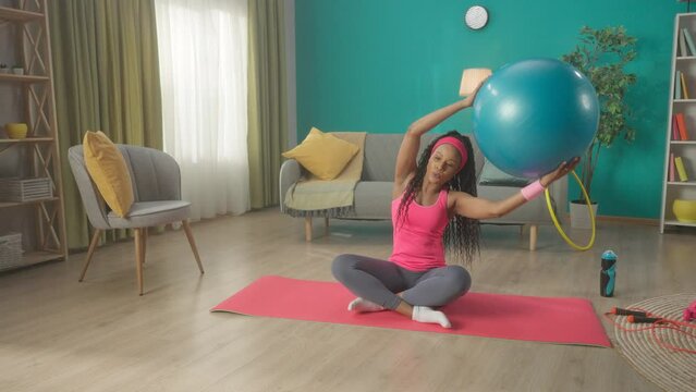 A young woman is sitting cross legged on a sports mat. African American woman raises straight arms with a fitness ball above her head and does side bends. HDR BT2020 HLG Material.