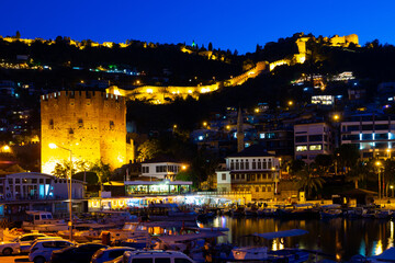 Illuminated pier of Alanya with moored boats in evening. View of Kizil Kule (Red Tower) and Castle of Alanya, Antalya Province, Turkey.