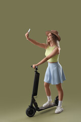 Young woman in summer hat with electric bike and mobile phone taking selfie on green background