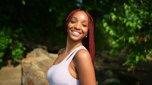 Outdoor portrait of natural Beautiful young African American woman long red braids hair style and perfect white teeth smile, posing in swimsuit at sunny summer day with green foliage beach background