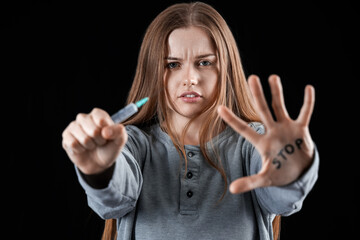 Young female junkie with syringe and word STOP written on her palm against dark background