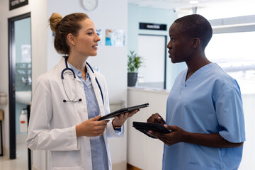Diverse female doctors using tablets and talking in corridor at hospital