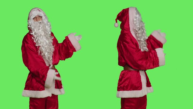 Father christmas advertising something, pointing aside while he stands against greenscreen backdrop with stanta claus festive costume. Man dressed like saint nick, xmas marketing campaign.