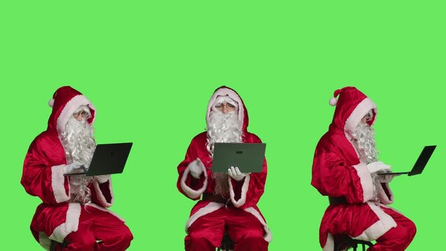Young man as santa claus using laptop, posing dressed in red and white festive suit during christmas holiday season. Saint nick sitting on chair scrolling on website, xmas celebration.