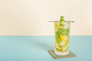 Glass of mojito with lemon, cucumber and ice cubes on blue table near white wall