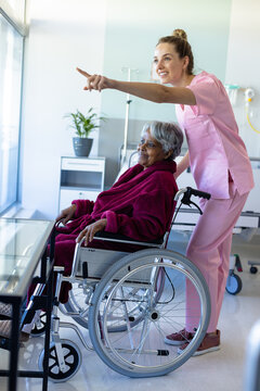 Diverse female doctor and senior female patient in wheelchair, looking through window at hospital