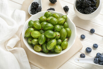 Bowl with fresh gooseberry, blackberry and blueberry on light wooden background, closeup