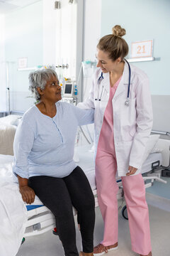 Diverse female doctor and senior female patient embracing and talking at hospital