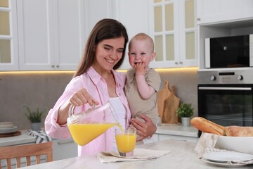 Obraz na płótnie Canvas Happy young woman holding her cute little baby while pouring juice into glass in kitchen