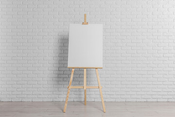 Wooden easel with blank canvas near white brick wall indoors