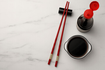 Bowl, bottle with soy sauce and chopsticks on white marble table, flat lay. Space for text