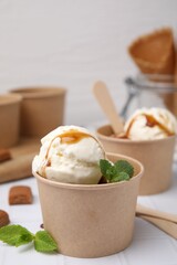 Scoops of ice cream with caramel sauce, mint leaves and candies on white tiled table, closeup