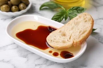 Bowl of organic balsamic vinegar with oil served with bread slice, basil and olives on white marble table, closeup