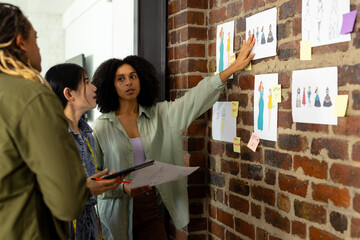 Diverse fashion designers using tablet and discussing design drawings on wall at studio
