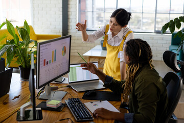 Diverse casual business colleagues using computer and laptop discussing work at desk in office