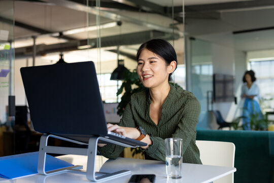 Happy asian casual businesswoman at desk in office using laptop on stand, smiling