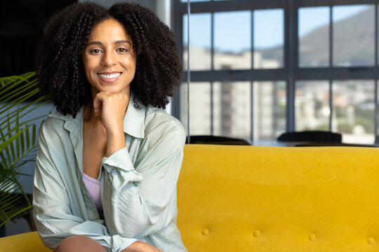 Portrait of smiling biracial casual businesswoman with curly dark hair on sofa in office, copy space