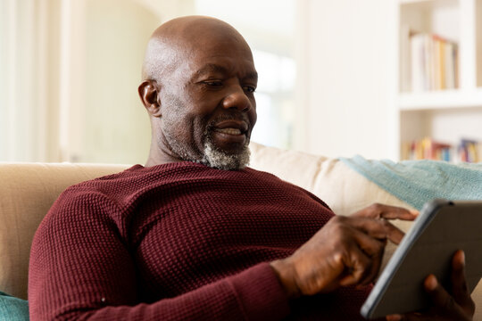 Happy african american senior man using tablet sitting in sunny living room - Powered by Adobe