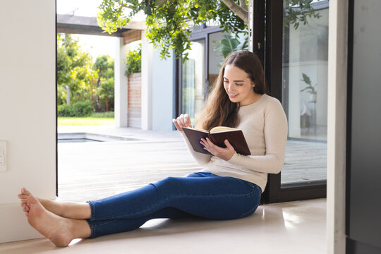 Happy caucasian woman sitting at wall and reading book at home