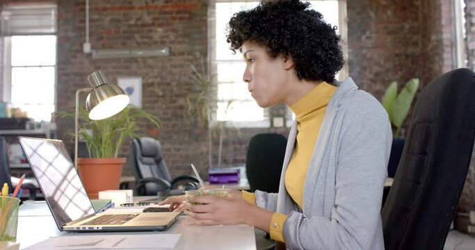 Focused biracial casual businesswoman using laptop and having lunch in office in slow motion