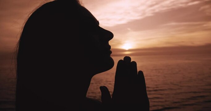 Close up young woman folded hands in prayer silhouette at sunset. Woman praying to God, standing outdoor on sea sunset background. Religion, spiritual and hope concept