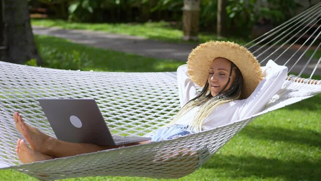 Glad woman freelancer digital nomad finished working on laptop during summer travel, lying in hammock. Woman freelancer satisfied with finish work on laptop computer and lies on hammock enjoying life.