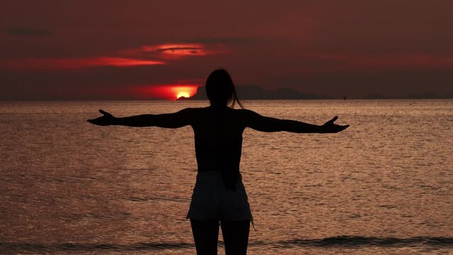 Rear view silhouette woman standing with her back to camera, looking at sun before sunset, enjoying beautiful seascape in the evening. Woman smoothly raises hands enjoying sea sunset while traveling
