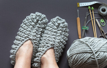 Top view of freshly knitted warm woolen house slippers on female feet and knitting accessories on dark gray background. Circular and straight knitting needles, crochet hook, scissors and skein of yarn