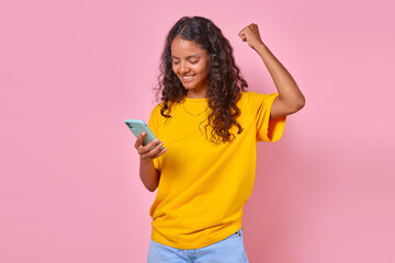 Young overjoyed ethnic Indian woman teenager holding phone and making victorious wave of hand using mobile application for online broadcast of sports match or lottery game stands on pink background