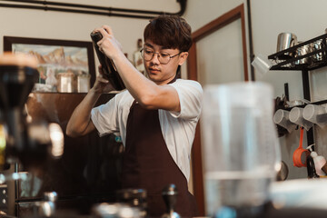 Barista Asian man holds in hands steel shaker at coffee shop. Asian man barista in white t-shirt and apron working at coffee shop. SME business coffee shop concept.